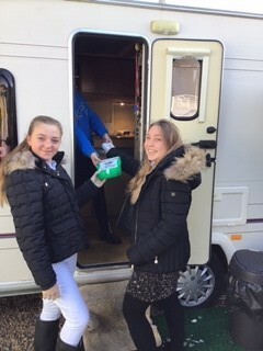 Two young Traveller girls stood outside a trailer handing over a first aid kit 