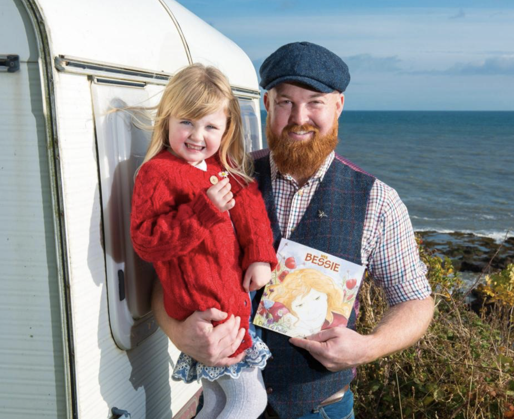David Pullar holding his daughter and his children book Wee Bessie. A white trailer to the left and the ocean to the right. 