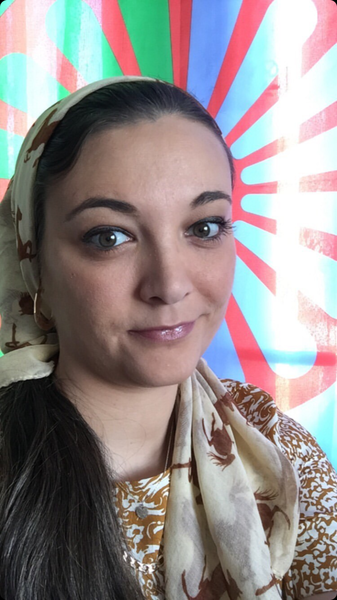 A photo of Nikki Hughes wearing a cream headscarf and gold earings smiling in front of the red and green Romani flag. 