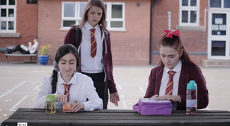 Two girls sat on a bench eating lunch and one girl stood behind looking mean at one of the girls 