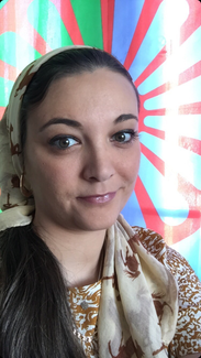 A photo of Nikki Hughes wearing a cream headscarf and gold earings smiling in front of the red and green Romani flag.