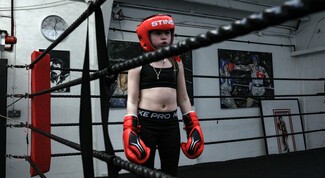 young white female wearing black sports clothes standing in a boxing ring with red gloves on