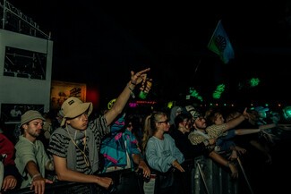 The row of young adults at the front of a crowd wearing festival clothes. The Romani Flag flies in the background