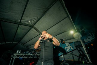 A white male, Sugar Shane, rapping into a mic and pointing with his finger. He is standing on a sheltered stage, it is night time.