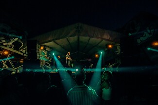 A night picture from the crowd watching a white male, Sugar Shane, performing on a stage. There are two blue overlapping stage lights.