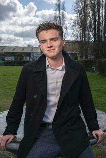 Jimmy Doherty wearing dark overcoat and white shirt leans against object in a field.