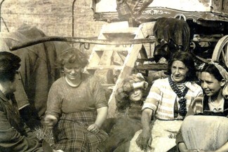 Romany Women sat outside of a wagon around a fire with a kettle on a prop in the middle 